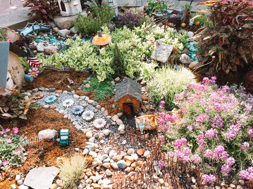 outdoor miniature garden with small sheds, stepping stones, rock pathway, and many leafy plants
